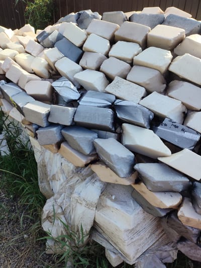 The scene depicts a large, irregularly stacked pile of diverse blocks situated outdoors in Prigorodnyi, Russia. The pile consists predominantly of two types of materials: numerous light-colored, off-white to cream-colored blocks with textured, porous, or slightly damaged surfaces, consistent with foamed insulation or lightweight construction material; and various grey-colored blocks that appear to be wrapped in a dark, fine-mesh netting. Some of these netted blocks exhibit tears or exposed underlying material. Interspersed are a few yellowish or beige blocks.
The blocks are arranged haphazardly, forming a substantial mound that extends from the foreground towards a dark brown, possibly wooden plank fence in the background. Green foliage, suggestive of bushes or small trees, is visible behind the fence. The ground surrounding the base of the pile consists of dark earth, scattered gravel, and patches of green grass. Crumpled white plastic sheeting is visible at the very bottom and around the base of the stack. No individuals, animals, or explicit actions are depicted. The lighting indicates an outdoor daytime setting with clear sunlight creating shadows on the blocks.