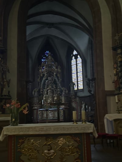 The image depicts the interior of a church in Vianden, Luxembourg. The focal point is a large, ornate altar with intricate carvings and golden embellishments. Behind the altar, a tall, multi-tiered structure with detailed ornamentation rises towards a vaulted ceiling. To the right of the altar, a stained-glass window with diamond-shaped panes in shades of yellow and blue is visible. In front of the altar, a white tablecloth covers a decorated surface. To the left of the altar, a vase with pink flowers is placed on a table. Various statues and decorative elements are scattered throughout the scene, including figures on pedestals and candelabras. The overall lighting is dim, creating a somber and reverent atmosphere.