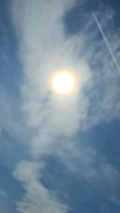 The image shows a bright sun with a halo effect, positioned in the upper center against a blue sky with scattered clouds. A contrail from an aircraft is visible in the upper right quadrant of the image, extending diagonally downwards. The scene appears to be an outdoor environment, likely during the daytime, with a focus on the sky and celestial phenomena. No people or man-made structures are discernible in this particular view, and the setting is generic enough that a specific location within a city like Таш-Асты, Russia cannot be determined.