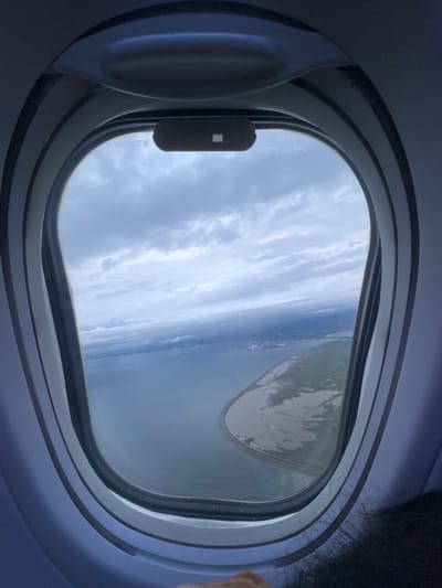 The image captures an aerial view from an airplane window, tilted to show the horizon at an angle. The oval window frame dominates the foreground, with the airplane's interior wall visible around it. Part of a person's dark hair is visible on the far right edge, suggesting a passenger is looking out.
Outside the window, a cloudy sky stretches above a large body of water, which appears to be a sea or extensive lake. A distinct strip of land is visible along the distant horizon, partially obscured by the clouds. Sunlight indicates it is daytime, and while clouds are present, visibility appears good. A reflection, possibly from the aircraft's interior or wing, is present on the right side of the window.
Several alphanumeric markings are visible on the window glass. On the lower-left side, oriented vertically, the text "S021632738" is clearly legible. Higher up on the left side, also vertically, "S-0216-223/69-L" is partially visible, likely representing part numbers or manufacturing codes for the window pane. The overall setting is an in-flight experience, observing the landscape from above.