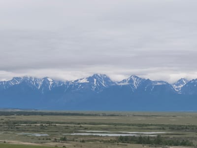 The image depicts a panoramic view of a mountainous landscape under a cloudy sky. In the foreground, a flat, marshy terrain is visible, dotted with small bodies of water and sparse vegetation. This expanse transitions into a more densely vegetated area with scattered trees, and further back, a wide, flat plain stretches towards the distant mountains. The mountains themselves are majestic and imposing, their peaks capped with snow and partially obscured by clouds. The overall impression is one of natural grandeur and serenity. The scene is believed to be located in the vicinity of Kharamodun, Russia, a region known for its dramatic natural beauty.