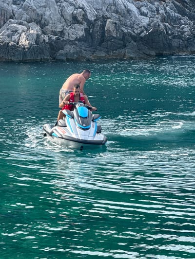 Two men are on a jet ski in the emerald green water off the coast of Dubraljevina, Montenegro. The man in front is driving the jet ski and wearing a red life vest. The man behind him is shirtless and wearing patterned shorts. In the background, a rocky cliff face rises from the water. The water is choppy, with white spray trailing behind the jet ski.