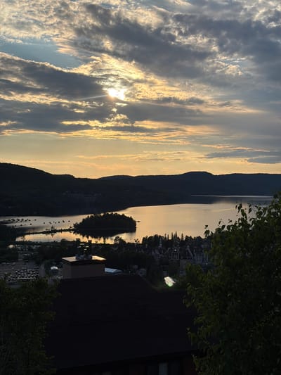 This is an aerial, high-angle view of a lake and surrounding landscape in Mont-Tremblant, Quebec, Canada, during sunset or sunrise. The sky is filled with dramatic clouds, with the sun partially obscured, casting a warm, golden light across the scene. A calm lake reflects the colors of the sky, with several small islands dotting its surface. On the far shore, dark, silhouetted mountains rise, with a cluster of buildings nestled at their base. In the foreground, the roofs of buildings and lush green foliage are visible. The image captures a serene and picturesque view of the natural beauty and the charming architecture of Mont-Tremblant.