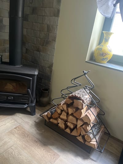 The image depicts an interior scene featuring a wood-burning stove and a decorative log holder. The stove is black and appears to be made of metal, with a glass door revealing a fire. Adjacent to the stove is a stone wall. In front of the stove, to the right, is a stack of firewood contained within a metal log holder shaped like a Christmas tree. A small decorative bird figurine is perched on top of the log holder. To the right of the log holder, on a windowsill, sits a yellow vase with blue and red decorative elements. The floor is covered in light-colored wood-look laminate flooring arranged in a herringbone pattern. The walls are painted a pale yellow. The overall setting suggests a cozy room, possibly a living room or dining area. There is no indication of the city of Староживотинное, Russia within the image itself.