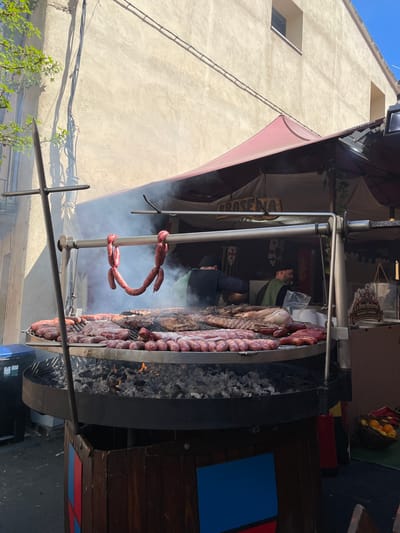 The image depicts a medieval-style food stall in Hostalric, Spain. Sausages are hanging on a metal pole. A large grill behind the pole is filled with various meats cooking over hot coals. Smoke rises from the grill and food. People are visible behind the stall's counter. Buildings are in the background.