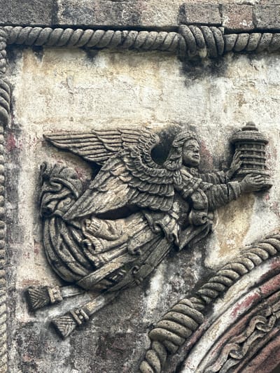 This is a close-up, eye-level shot of a stone carving depicting an angel with wings, holding a lantern. The carving is part of the facade of the San Miguel Arcángel church in Huejotzingo, Mexico. The angel, rendered in grey stone, has detailed feathered wings, flowing robes, and a serene expression. It is positioned in the center of the frame, with its left hand holding a multi-tiered, cylindrical lantern. The stone is weathered and shows signs of age, with patches of discoloration and texture variations. The background consists of the rough, textured stone wall of the church, adorned with rope-like decorative elements and other carved architectural details, some of which show traces of red pigment. The lighting appears to be natural daylight, casting subtle shadows that highlight the relief of the carving.