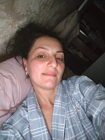 The image shows a woman lying on a pillow, facing the camera. She has dark, curly hair and is wearing a gray and white plaid robe. Her left ear is visible, adorned with a hoop earring. The background appears to be an interior space with a textured wall on the left and a dark, cluttered shelf or cupboard on the right. The lighting is dim. No specific details indicating a location in Tsandrypsh, Abkhazia, are visible in the image.
