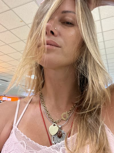 A medium shot captures a Caucasian woman with blonde hair, from the chest up, indoors. She is wearing a pink lace camisole and several necklaces, including a gold chain with a heart pendant and a silver pendant with an intricate design. A subtle nose stud is visible on her right nostril. Her left hand is raised and touching her head, obscuring part of her face with her hair. The background consists of a tiled ceiling and a glimpse of an orange sign in the distance, suggesting an airport terminal or similar public building. The lighting is bright and diffused. The scene appears to be an interior space within the Katunayake airport, Sri Lanka.