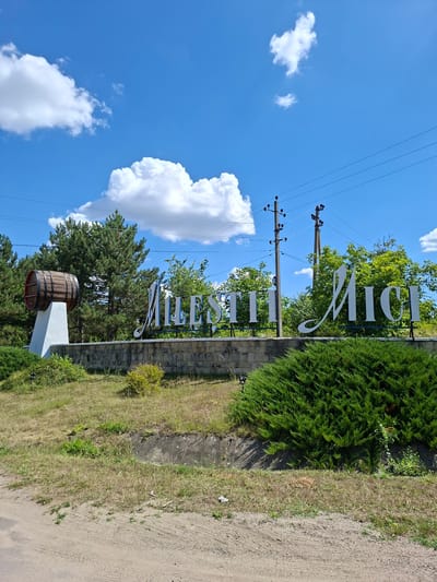 In Mileștii Mici, Moldova, a sign with large white, stylized lettering on a dark blue background announces the name of the town. The sign is situated behind a stone wall and in front of a dense evergreen bush. To the left of the sign, a large wooden barrel rests on a white pedestal. In the background, several tall pine trees are visible, along with other deciduous trees and shrubs. Overhead, a bright blue sky is dotted with fluffy white clouds. Electrical poles and wires are present above the trees. The foreground consists of a dirt road or path and dry grass. The overall scene depicts an entrance sign for the town.