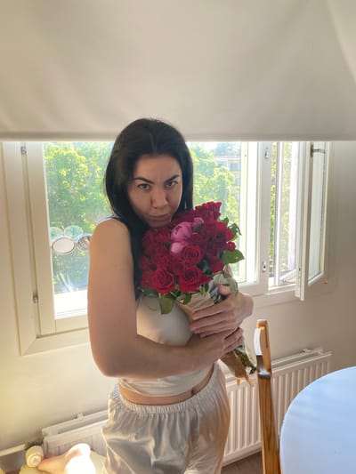 A woman with dark hair stands indoors, holding a bouquet of red roses and pink peonies. She is wearing a white sleeveless top and white satin pajama bottoms. She is positioned in front of a window that overlooks a tree-lined street and buildings, suggesting an urban environment. The window has white frames and is partially open. A radiator is visible below the window, and a wooden chair is to the right of the woman. The lighting indicates it is daytime, possibly late afternoon, with sunlight streaming in. The overall scene appears to be a domestic setting, possibly an apartment in Kotka, Finland, given the architectural style of the window and radiator, and the appearance of the surrounding greenery.