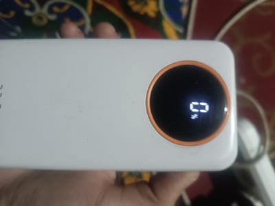 A hand holds a white rectangular power bank. The power bank has a circular black screen with an orange ring around it. The screen displays "0%" in white, indicating a zero percent battery charge. The background is blurred, showing a patterned surface with red and green hues.
