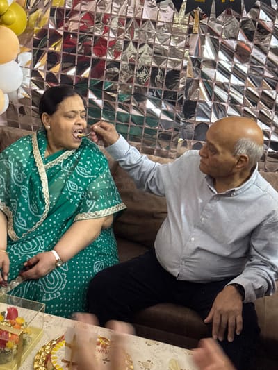 In Bidhannagar, India, an indoor scene features two older adults seated on a brown sofa. An older man, bald with gray hair on the sides, wearing a light blue long-sleeved shirt and dark trousers, is extending his right arm to feed a spoonful of light-colored food to an older woman seated to his left. The woman, dressed in a green patterned sari with cream-gold trim, has her mouth open to receive the food and her eyes partially closed. She wears a white wristwatch on her left wrist and has a reddish decorative application on her right hand. The background wall is covered with numerous small, reflective silver square panels. Yellow, orange, and white balloons are visible in the upper left corner. In the foreground, on a light-colored table, a gold-colored scalloped tray holds a partially visible cake or dessert, and a clear acrylic box contains decorative items.