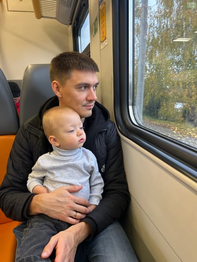 An adult male holds a baby on his lap inside a train car, located in the vicinity of Пески, Russia. The man, with short brown hair, a light mustache, and a nose piercing on his left nostril, is wearing a black puffer jacket over a dark hooded garment and blue jeans, with a gold band on his left ring finger. The baby, appearing to be an infant, has light hair and is dressed in a light grey long-sleeved turtleneck and dark grey pants. Both individuals are looking out the train window towards the right side of the frame. Through the window, an autumnal landscape is visible, featuring trees with yellow and orange foliage, and a blurred view of what appears to be a road or path and a distant light-colored structure. The train interior features light-colored walls, grey seatbacks, an orange-brown seat cushion, and various safety or informational signs with Cyrillic text affixed near the window. A partial view of another person wearing a black beanie and red clothing is visible in the background.