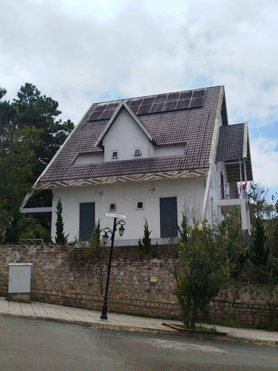 The image depicts a white, two-story house with a dark tiled roof, a portion of which is covered with solar panels. The house is situated on a slight incline, behind a stone and brick wall. A paved sidewalk runs along the front of the wall, adjacent to a street. A black lamppost with two lights and a solar panel is positioned on the sidewalk in front of the house. Numerous trees and bushes surround the property. In the background, the sky is partly cloudy. The scene appears to be in a semi-rural or suburban area, consistent with the landscape of Lang Biang - Da Lat, Vietnam, known for its hilly terrain and vegetation. No people are visible in the image.