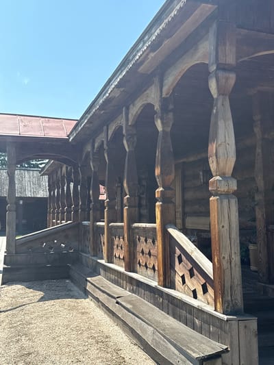 The image depicts the exterior of a traditional wooden log house with a covered porch, likely a historical exhibit or museum in Shushenskoye, Russia. The porch is supported by elaborately carved wooden posts, each with decorative turning and a hexagonal base. A wooden railing with a diamond-shaped cutout pattern runs along the porch. A wooden bench is situated in front of the porch railing. The ground in front of the porch is covered with light-colored gravel. The log walls of the house are visible behind the porch. The sky is clear and blue with a few wispy clouds. The overall scene suggests a warm, sunny day.