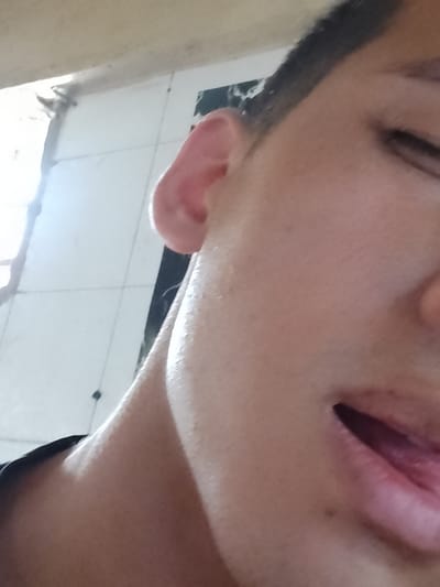 The image presents a close-up, angled view of the right side of a person's face, likely taken from a lower perspective. The subject's skin is light-toned, and parts of the right ear, eyebrow, closed or squinting eye, nose, and mouth are visible. The mouth is open, revealing a portion of the tongue, suggesting an action like tasting, a playful expression, or yawning. Bright light, possibly from a window or overhead source, illuminates the scene, creating strong highlights on the person's skin, especially on the cheek and neck, and casting soft shadows.
The background consists of light-colored, possibly tiled surfaces, with visible grout lines or structural separations. There are also darker, blurred elements in the background, suggesting architectural features or other objects out of focus. The bright lighting indicates that the photo was likely taken during the daytime in an indoor or semi-enclosed setting. No specific objects other than the person's body parts are clearly identifiable. There is no visible text in the image. The scene is situated in El Dátil, Venezuela.