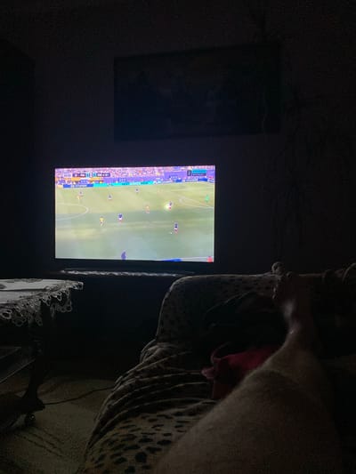 In a dimly lit room in Zastavna, Ukraine, a large flat-screen television displays a live soccer match. The score shows France 0 and Sweden 0 at 43:55. A person's legs and feet are visible in the foreground, draped in a leopard-print blanket, indicating they are likely sitting or lying down and watching the game. A small table with an ornate lace tablecloth is positioned to the left of the television. The room appears to be a living space, with the television being the central focus of attention.