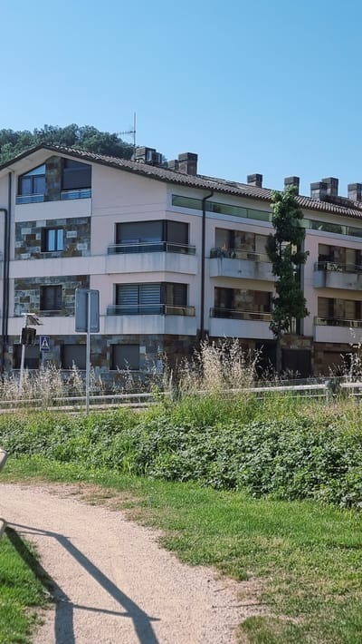 The image depicts a modern apartment building in Sant Joan les Fonts, Spain. The building has multiple stories, with balconies on each level featuring glass railings. The facade is a combination of light-colored plaster and stone cladding. Several windows with dark shutters are visible, as well as some air conditioning units on the roof. A tall, slender tree stands in front of the building. To the left of the building, a traffic signal and a pedestrian crossing sign are present. The foreground shows a gravel path running alongside a lush green area with tall grasses and bushes, indicating a natural or park-like setting. The sky is clear and blue.