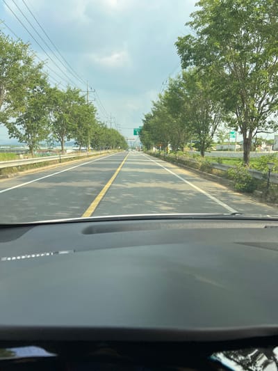 The image is a first-person view from inside a vehicle, looking forward through the windshield. The vehicle is on a paved road lined with trees and utility poles. The road has a yellow center line and a white lane line. In the distance, there is signage indicating a turn-off. The sky is partly cloudy. The scene appears to be in a rural or suburban area of Nonsan-si, South Korea.