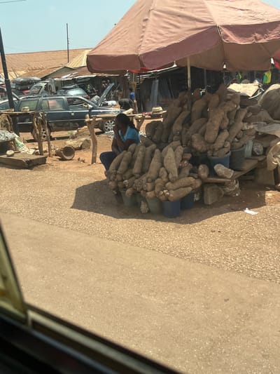 The image depicts an outdoor market scene in Karshi, Nigeria, under bright daylight. The main subject is a large, tall pile of yams or similar root vegetables, prominently displayed on the ground to the right of a paved area. A person wearing a blue top is seen sitting or crouching beside the yam pile, likely a vendor tending to their goods.
To the left, a large, reddish-brown market umbrella provides shade, extending over an unseen portion of the market. In the background, beyond the yam stall and umbrella, several vehicles are visible, some appearing to be parked or undergoing repair, with open hoods and visible car parts. Other people can be seen in the mid-ground, moving among the cars and what appear to be other market stalls or structures.
The ground is a mix of light-colored, speckled pavement and dusty earth. Strong shadows are cast by the market umbrella and other objects, indicating a sunny day, likely during the late morning or early afternoon. The setting suggests a bustling commercial area, characteristic of a local market in West Africa. No discernible text is visible within the image.