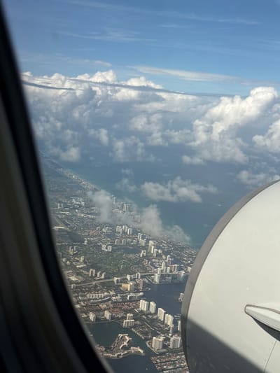 View from an airplane window over North Miami Beach, United States of America. Visible are a white airplane wing flap, sky with cumulus clouds, coastline, intracoastal waterway, residential and commercial buildings, and green spaces.
