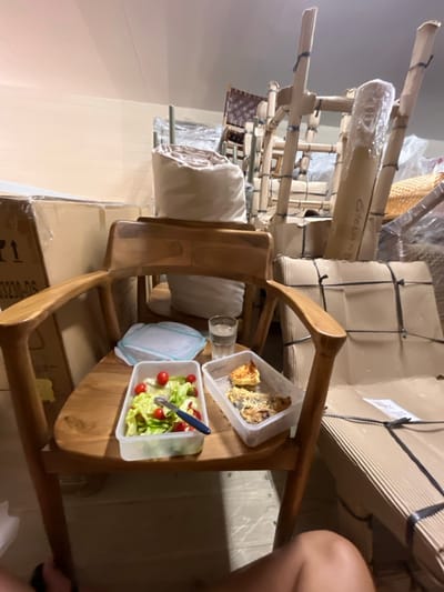 A wooden chair with a rounded seat holds a container of salad and a container of baked goods. A glass of clear liquid sits nearby. The chair is surrounded by stacked, packaged furniture, including chairs with woven seats and others wrapped in plastic. The scene has a cluttered, storage-like feel. A person's leg is visible in the background, suggesting someone is nearby.