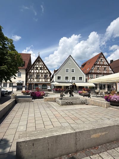 The image depicts a town square in Ebermannstadt, Germany, on a clear day with scattered clouds. In the foreground, a paved square with large square tiles leads towards a central water fountain adorned with bronze sculptures of animals, possibly pigs, and a figure with a barrel. Planters overflowing with colorful flowers surround the fountain.
Several buildings line the square. On the left, a light-colored building with a white facade and dark-framed windows is visible. Adjacent to it is a traditional half-timbered building with a dark timber frame and white infill, featuring multiple windows and a gabled roof. Further to the right, a modern, light gray building with a simple gabled roof stands next to another half-timbered structure with a red tile roof. Outdoor seating areas with tables and chairs are set up in front of some of the establishments, covered by large cream-colored umbrellas.
A white car is parked on the left side of the square. A person in a light-colored shirt can be seen standing near the outdoor seating on the right. The overall scene suggests a pleasant, public space in a German town.