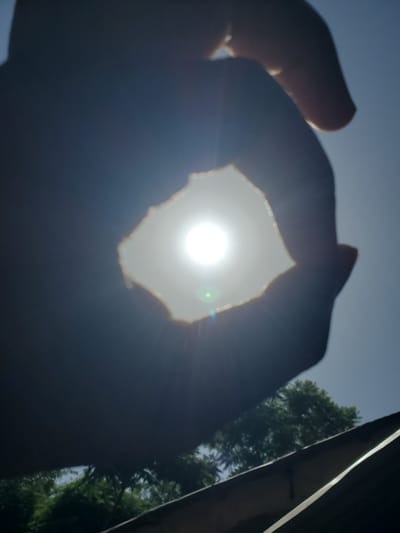 The image captures a close-up, low-angle view of a person's hands silhouetted against a bright sun. The sun is positioned in the center, peeking through a roughly torn or shaped opening between the fingers. The light from the sun is intense, creating a halo effect and lens flare. The hands are dark and indistinct, with only the outlines and the rough edges of the opening clearly visible.

In the background, a clear blue sky is visible above the sun. To the right, the dark, blurred shape of a building or structure runs vertically along the edge of the frame, with a bright sliver of light reflecting off its surface. Behind this structure, and slightly to the left, are the dark green shapes of trees, suggesting an outdoor setting. The overall impression is of a bright, sunny day. The time of day appears to be mid-day, given the high position of the sun. The scene is likely set outdoors, possibly in a residential or natural area given the presence of trees. The act of holding the hands up to frame the sun could suggest a moment of contemplation, playfulness, or simply an attempt to shield the eyes from the glare. No text is visible in the image. The context provided, "Kabwir, Nigeria," places this scene in that specific geographical location.