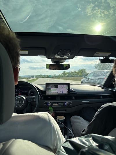 A view from inside a car, possibly a 2016-2024 Audi A4 or A5, shows the driver's perspective. The car is traveling on a highway under a partly cloudy sky. The GPS navigation display shows that the car is approximately 57 km from Exit 203, leading to Svitavy, suggesting the location is not Hulín. The driver is partially visible, with only the top of their head and sunglasses visible. The passenger seat contains a white sweater or blanket, a coffee cup, and a bottle of Turkish cranberry juice, "BARK." The speedometer reads 131 km/h, and the total distance is 1606.9 km. There's another car in the left lane.