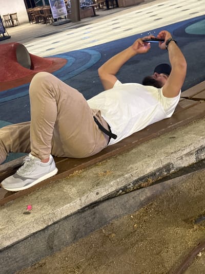 A man is captured from the side, mid-shot, as he sits on a ledge and uses his smartphone. He is wearing a white t-shirt, khaki pants, and light grey athletic shoes. A black watch is visible on his left wrist. His right arm is bent, holding the phone up in front of him, and his gaze is directed at the screen. He has a beard and is wearing a dark baseball cap. The background includes a colorful, abstract pattern on the ground, possibly a mural or pavement design, with shades of blue and white. To the right of the man, the ground transitions to a rough, dirt-like texture with scattered debris. The setting appears to be outdoors, possibly a public space, with hints of seating areas and signage visible in the blurred background. The lighting suggests it might be evening or late afternoon, with artificial light casting shadows.