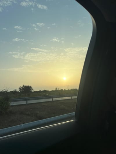 An exterior scene is depicted from the interior of a vehicle, looking out a side window during what appears to be sunrise or sunset. The sun, a bright, circular disc, is positioned low on the horizon slightly to the right of the frame's center, casting a golden-yellow light across the sky. The upper sky is light blue, transitioning to a warm yellow-orange closer to the horizon, with scattered, small white cloud formations visible throughout. In the midground, a dark asphalt road or highway is visible, running horizontally across the lower portion of the image, bordered by a metallic guardrail on its left side. Beyond the road, the landscape consists of a dense, dark green, mostly level terrain with various trees and shrubs. A faint, dark silhouette of what appears to be an electrical transmission tower is visible on the horizon, slightly to the right of the sun. The immediate foreground is occupied by the dark, curved interior frame of the vehicle's window and potentially a portion of the door panel, obscuring the right and top edges of the view. The scene is located in a rural or semi-rural area near Alcácer do Sal, Portugal. No people are visible.