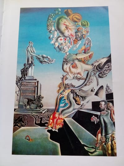 This is a surrealist painting depicting a dreamlike landscape. In the foreground, a man in a dark suit and tattered lower garments is embraced by another figure, who is holding a severed head. They are standing on a dark, angular surface. To the left, a monument displays the words "GRAMME," "CENTIGRAMME," and "MILLIGRAMME." A lion statue stands beside it. A distorted human form with a melting face is prominent in the upper right. Numerous abstract and organic shapes float in the blue sky. There is no discernible location within the city of Zhmerynka, Ukraine, in this artwork.