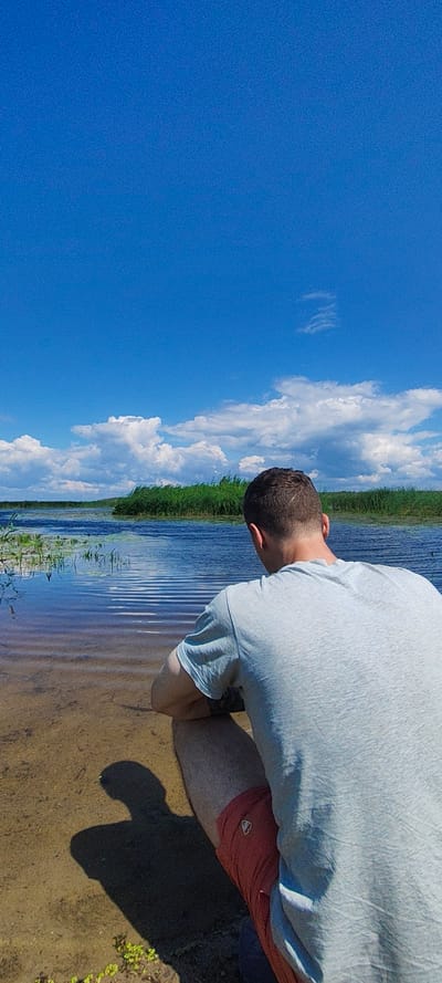 A person is crouching by the edge of a body of water, facing away from the camera. The person is wearing a light blue t-shirt and red shorts. Their shadow is cast on the sandy ground. The water is calm with ripples reflecting the sky. In the background, there is a dense line of green reeds or tall grass bordering the water, and beyond that, a faint treeline is visible. The sky is a vibrant blue with scattered white clouds. The scene is set in a natural environment, likely a lake or riverbank in the city of Malaya Listovka, Russia, on a clear day.