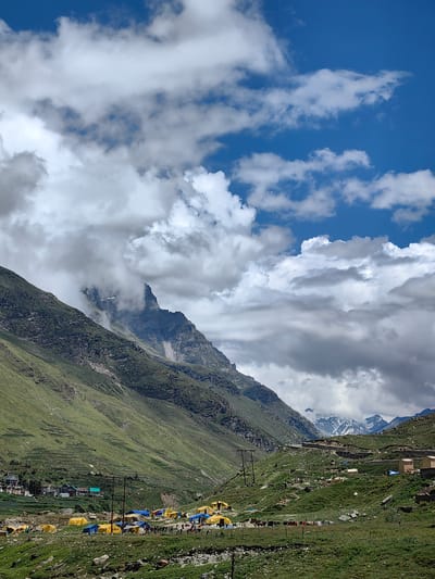The image depicts a mountainous landscape in the region of Koksar, India. A valley floor is populated with numerous tents, predominantly yellow and blue, indicating a campsite or temporary settlement. Some buildings are visible in the background, nestled on the slopes of the surrounding mountains. Power lines and poles are present throughout the scene, suggesting the presence of infrastructure. The sky is partially covered with dramatic, grey and white clouds, with patches of bright blue visible. The mountains themselves are steep and covered with green vegetation, with some rocky outcrops and snow-capped peaks visible in the distance. A winding road can be seen traversing the mountainside. The overall scene conveys a sense of a remote, high-altitude environment with human activity in the form of camping and settlement.