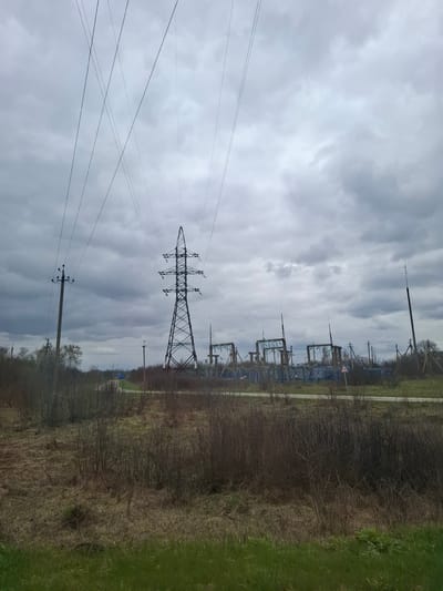 This image, rotated 90 degrees clockwise, depicts an industrial landscape dominated by electrical infrastructure under an overcast sky. In the foreground, multiple power lines crisscross the frame, emanating from several utility poles and a large lattice-work electricity pylon situated towards the left-center. Further in the background on the right, an electrical substation with numerous transformers and smaller pylons is visible.

To the right of the substation, there's a patch of green grass, and extending across the mid-ground, a wide strip of barren trees and bushes with a dirt path or road running through it. The bare branches suggest late winter or early spring. A red-bordered triangular road sign with a black 'S' shape on a white background, indicating a winding road ahead, can be seen near the substation. The sky is filled with grey clouds, creating a diffused light across the scene, characteristic of a cloudy day. The setting appears to be a rural or semi-industrial area near Panikovets, Russia, dedicated to power transmission. No people or specific activities are observed.