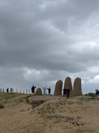 The image displays a scene at a sandy beach featuring the iconic "La Mano" (The Hand) sculpture in Punta del Este, Uruguay, under an overcast sky. The photograph is oriented vertically, requiring a 90-degree counter-clockwise rotation to be viewed correctly.
The central subject is the large, concrete sculpture depicting five human fingers partially submerged in the sand. Several people are present around the landmark. A few individuals are seen standing on a wooden walkway that runs parallel to the base of the sculpture. One person near the lower fingers appears to be posing with outstretched arms, while others are observing or walking. Further along the walkway, more people are visible, some standing closer to the sculpture's middle fingers. In the background, on a higher sandy dune, additional people are observing the scene.
The setting is a beach environment characterized by light brown sand and sparse green vegetation growing on the dunes. A wooden fence or barrier is partially visible along the edge of the sandy path. A row of small, colorful flags is positioned along the left side of the sandy area. The weather is overcast, with thick, grey clouds dominating the sky, indicating a cloudy daytime. No visible text is discernible in the image. The overall impression is one of a popular tourist spot, with visitors leisurely exploring and interacting with the unique artwork.