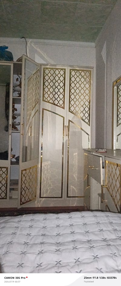The image depicts an interior scene, likely a bedroom or dressing area, with a prominent wardrobe and a quilted bed in the foreground. The wardrobe is a tall, white structure with ornate gold detailing in a lattice pattern on its doors. It appears to be a built-in unit, with shelves visible in an open section on the left side, displaying decorative items. To the right of the wardrobe is a smaller cabinet or dresser, also with gold accents and an open drawer revealing its contents. The bed is covered with a white quilted comforter featuring a repeating cross-like pattern. The lighting suggests it is daytime, with some light reflecting off the wardrobe's surfaces. The camera information at the bottom indicates the photo was taken with a CAMON 30S Pro at 23mm focal length, f/1.8 aperture, 1/20s shutter speed, and ISO 3784. The location is identified as Fayzabad, Kalot, Tajikistan. No people are visible in the image.
