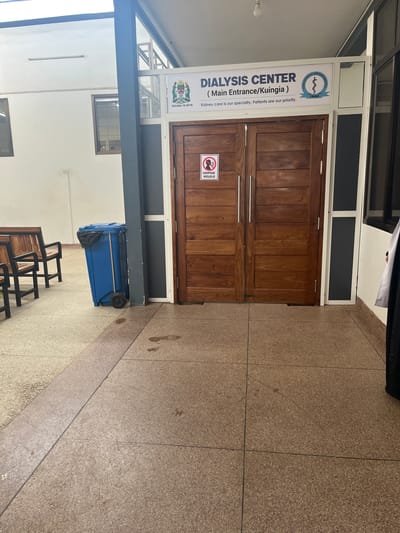 The image depicts the entrance to a dialysis center at TRRH (possibly Tumaini Regional Referral Hospital). The entrance is through a pair of wooden doors with a sign indicating that it is the "Main Entrance/Kuingia" and identifies the facility as a dialysis center. The sign also states, "Kidney care is our specialty. Patients are our priority". The sign includes the emblem of the Tanzanian Ministry of Health (Wizara Ya Afya). Another sign is seen saying "Usipige Kelele" which means "Do not make noise". A blue wheeled garbage bin is located near the entrance. Some wooden chairs are visible in the foreground. The floor is tiled with beige stone, and there are footprints on the floor.