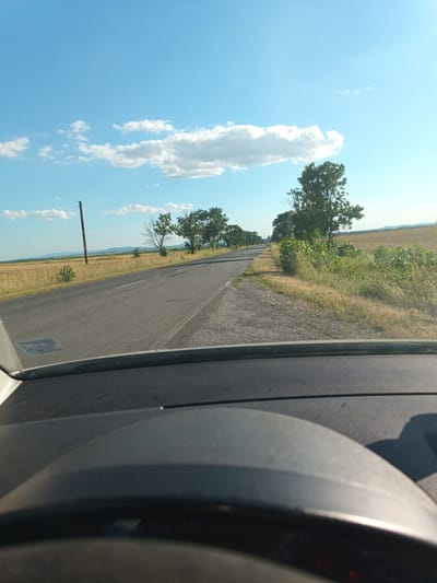 The image shows a view from inside a car, looking forward and slightly to the right. The foreground is dominated by the dark, curved dashboard and the rim of the steering wheel. The road ahead is asphalt and curves gently to the left, with a gravel shoulder on the right. Fields of dry grass and crops stretch out on either side of the road, with scattered trees and bushes. A utility pole is visible on the left side of the road. In the distance, rolling hills can be seen under a bright blue sky with scattered white clouds. The scene is consistent with a rural road in the vicinity of Okop, Bulgaria, likely during the daytime in a warm season.