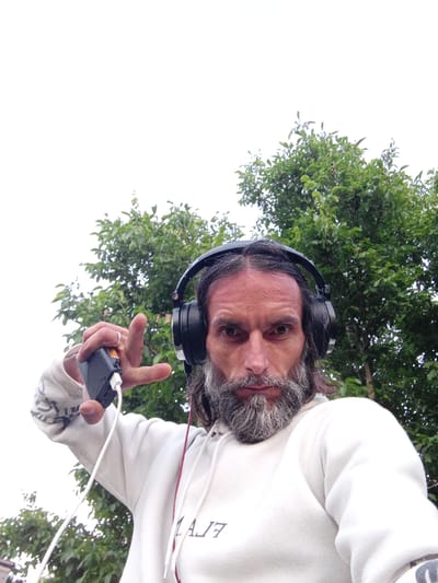 A man with long, dark hair and a gray beard, wearing black headphones and a white hoodie with a design on the front, holds a black rectangular device with a white wire attached in his left hand, his index finger pointing upward. He is outdoors, with green trees and a bright sky visible in the background. The man's face is partially obscured by his beard and headphones, but his eyes are visible, looking directly at the camera. A ring is visible on his left ring finger. The scene appears to be in a rural or suburban outdoor setting, possibly within or near the city of Голубинское сельское поселение, Ukraine.