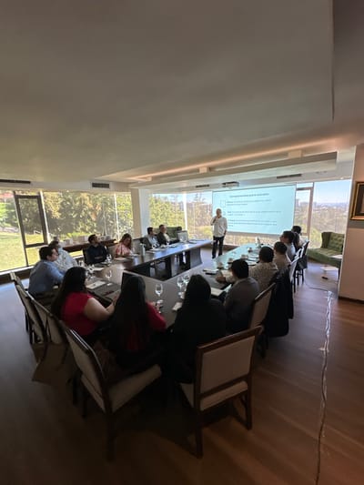 A group of people is seated at a long table in a bright room, attending a presentation. A man stands near a projected screen displaying text in Spanish. The room has large windows offering a view of trees and greenery. Water glasses are placed in front of each attendee. Some people are facing the presenter, while others appear to be looking at their phones or talking. The room has a modern design with a neutral color palette.