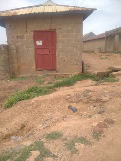 The image depicts an outdoor scene in Goni Gara, Nigeria, under an overcast sky, suggesting a cloudy or possibly rainy day. The primary focus is a small, single-story building constructed from concrete blocks, with a corrugated metal roof that appears weathered. A red metal door, divided into panels, is set into the front of the building. Above the door, a small, rectangular white sign is affixed to the wall. The building has the marking "H48" written in black on its left side.
The immediate surroundings are characterized by rough, sandy terrain with patches of green grass growing sporadically. In the foreground, the ground is uneven and appears to be a mixture of dirt and rocks. There are several pieces of debris scattered on the ground, including plastic, and what might be animal waste. A small, rudimentary drainage channel is visible to the right of the building, with some debris lodged within it.
In the background, other structures are partially visible, also appearing to be simple buildings with corrugated metal roofs. The overall impression is of a rural or semi-urban environment, likely a residential or small commercial area, characterized by unpaved ground and basic construction. No people are visible in the frame, and the scene appears still, with no discernible activity. The lighting is diffused, consistent with a cloudy day. The visible text "H48" and the white sign on the door are the only identifiable textual elements.
