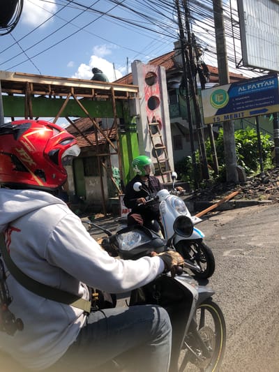 In Banjarkemantren, Indonesia, a motorcycle passenger wearing a red helmet and a grey hoodie rides behind another motorcyclist who is wearing a green helmet and a dark jacket. Both are on scooters. Behind them, a building is under construction, with scaffolding and construction materials visible. A signpost advertising "AL FATTAH" Islamic boarding school, along with its address and other information, stands prominently on the right side of the road. The sky is clear with some clouds, and power lines are strung across the top of the image.