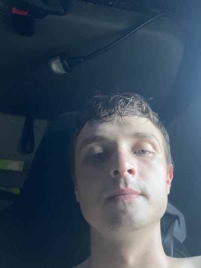 The media file is a close-up, tilted selfie of a young Caucasian person with wet hair and a pale complexion. The lighting is dim, suggesting it might be nighttime or an indoor setting with low light. The person's head is angled, with their face partially obscured by their hair. Their eyes are open, and they appear to be looking towards the camera. Their lips are slightly parted. The background is dark and appears to be the interior of a vehicle, possibly a car or truck, with a dark fabric seat or headrest visible. There's a black cable with a small, illuminated object at the end, possibly a light or accessory, visible on the left side of the frame. The overall mood is casual and personal. There is no visible text in the image.