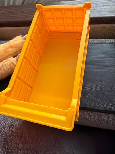 An overhead shot shows an empty, bright orange plastic storage bin with reinforced corners. The bin is placed on a textured grey surface that appears to be upholstery. To the left of the bin, a portion of a fluffy, light brown animal's paw is visible, suggesting the animal is resting nearby. The background consists of dark, textured surfaces, possibly furniture. The scene is indoors and well-lit. The city of Mirnoye, Ukraine, is not depicted in this image.