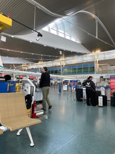 The image captures a scene inside an airport terminal, likely located in Tambon Mai Khao, Thailand, given the presence of signage in Thai. The environment features a high, modern ceiling with exposed metal beams and light fixtures. The floor is a speckled, greenish-blue tile.

In the foreground, to the left, a light brown woven bench seat with white metal legs is visible. Next to it, a red fire extinguisher stands upright.

The middle ground shows a man in khaki pants and a black long-sleeved shirt pushing a white baggage cart with a partially visible black bag. He is walking away from the camera. Further back, to the right, two individuals stand near a row of check-in counters. One, wearing a black hoodie with "MADHAPPY" and a Yankees logo, stands beside several black suitcases on a trolley. The other person, dressed in dark clothing, is partially obscured.

The background reveals multiple check-in counters labeled with numbers (e.g., F02, F03, F04, F05, F06, F09) and some signage, including what appears to be "AEROFLOT Russian Airlines" and "BUSINESS CLASS" in both English and Russian. Other signs in Thai are also present. The overall impression is a busy airport environment with people engaged in travel-related activities.