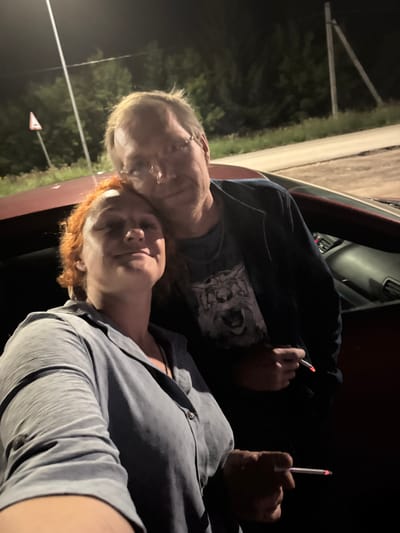 Two individuals, a man and a woman, are posing for a selfie outdoors at night. The woman is in the foreground on the left, her head tilted upwards and a slight smile on her face. She has red hair and is wearing a light blue collared shirt. The man is behind her and to the right, with his head leaning on hers. He has short, light-colored hair and is wearing glasses and a dark blue jacket over a t-shirt with a graphic print of a roaring animal. Both individuals are holding lit cigarettes. They are positioned next to a red car. The background shows a dimly lit road with trees and power poles. There is a triangular warning sign visible in the distance to the left. The overall atmosphere suggests a casual, late-night roadside stop. The location is identified as Kutlu-Bukash, Russia.