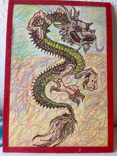 The image is a photograph of a drawing on a wooden board, framed by a red border. The drawing depicts a stylized dragon with green scales, spiky brown armor along its back, and tan limbs. The dragon's head is detailed with sharp teeth, red eyes, and a long, white mustache-like appendage. Its body is coiled in an S-shape. The background of the drawing is a chaotic swirl of yellow, blue, and red lines. The wooden board itself appears to be placed on a white surface, and a textured white wall is visible in the upper left corner. There are no people or discernible actions in the image. The scene does not provide any information about its location within Schwäbisch Hall, Germany.