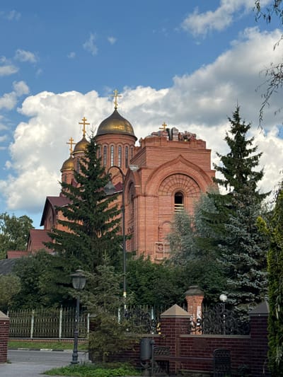 In Il'inskiy, Russia, a large brick church with golden domes dominates the scene under a partly cloudy blue sky. The church features multiple domes, each topped with a golden cross, and is constructed from red brick. A tall, dark green evergreen tree stands in front of the church, partially obscuring it. A lamppost with a dark metal frame and a glass lantern is positioned to the left of the tree. The foreground shows a paved area with a grassy verge and a dark metal fence. The overall impression is of a peaceful, imposing religious structure set amidst lush greenery.