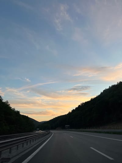 The image depicts a highway at dusk, cutting through a forested valley. The sky is a gradient of pale blue to soft orange and pink hues, with wispy clouds. Dense, dark green trees line both sides of the highway, creating a sense of enclosure. The road itself is paved asphalt with white lane markings. A silver guardrail is visible on the left side of the road. In the distance, a single car with its headlights on is driving away from the camera. A sign that reads "BETWILD" is visible on the right side of the highway. The scene conveys a sense of travel and the transition from day to night. The location is identified as the highway near Churek, Bulgaria.